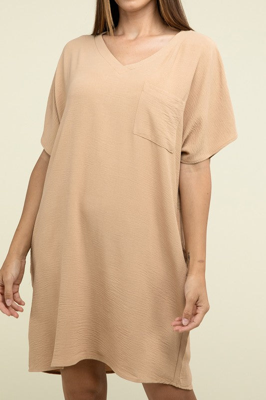 A woman wearing a loose light brown Woven Airflow V Neck T-Shirt Dress with Pockets stands against a plain background. Made from breathable fabric, this summer dress includes pockets where her hands rest as she smiles slightly.