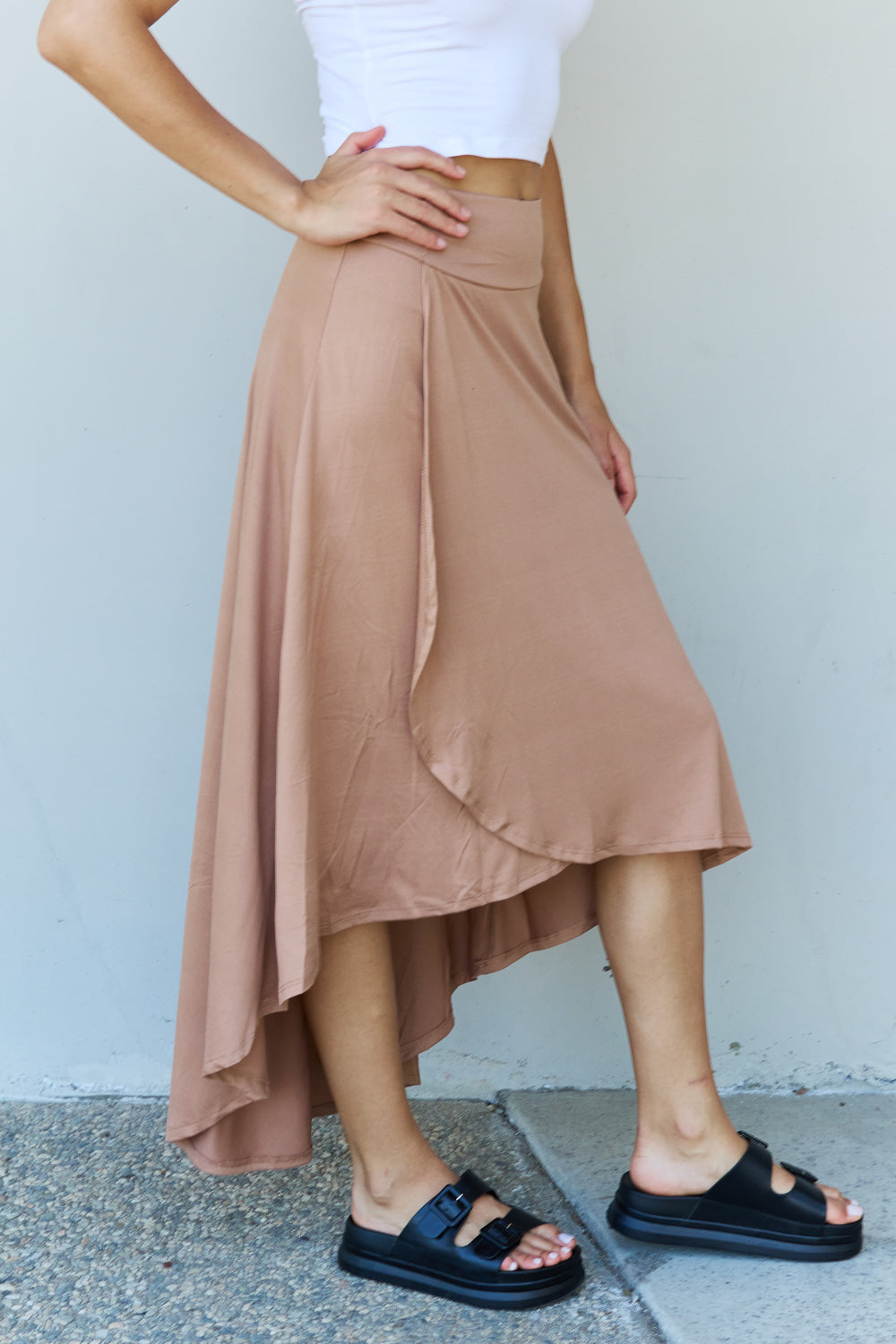 Person wearing a white crop top, the Ninexis First Choice High Waisted Flare Maxi Skirt in Camel with an asymmetrical hemline, and black sandals, standing against a light-colored wall.