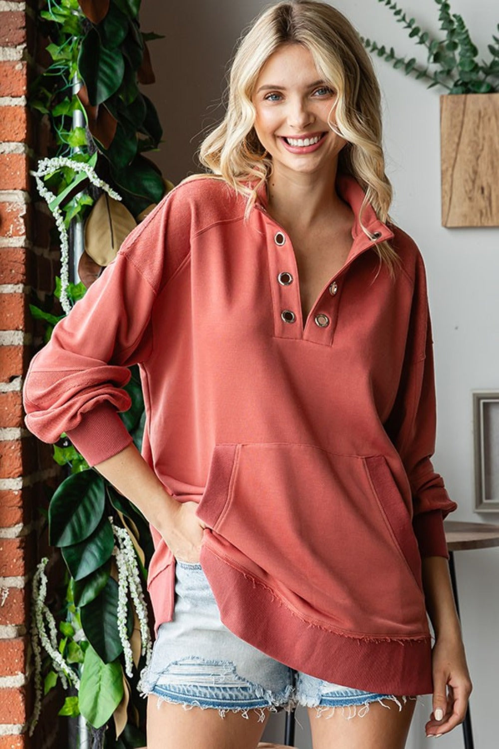 A woman with blonde hair is wearing a First Love Grommet Split Neck Kangaroo Pocket Sweatshirt in coral, standing indoors near a brick wall and plants.