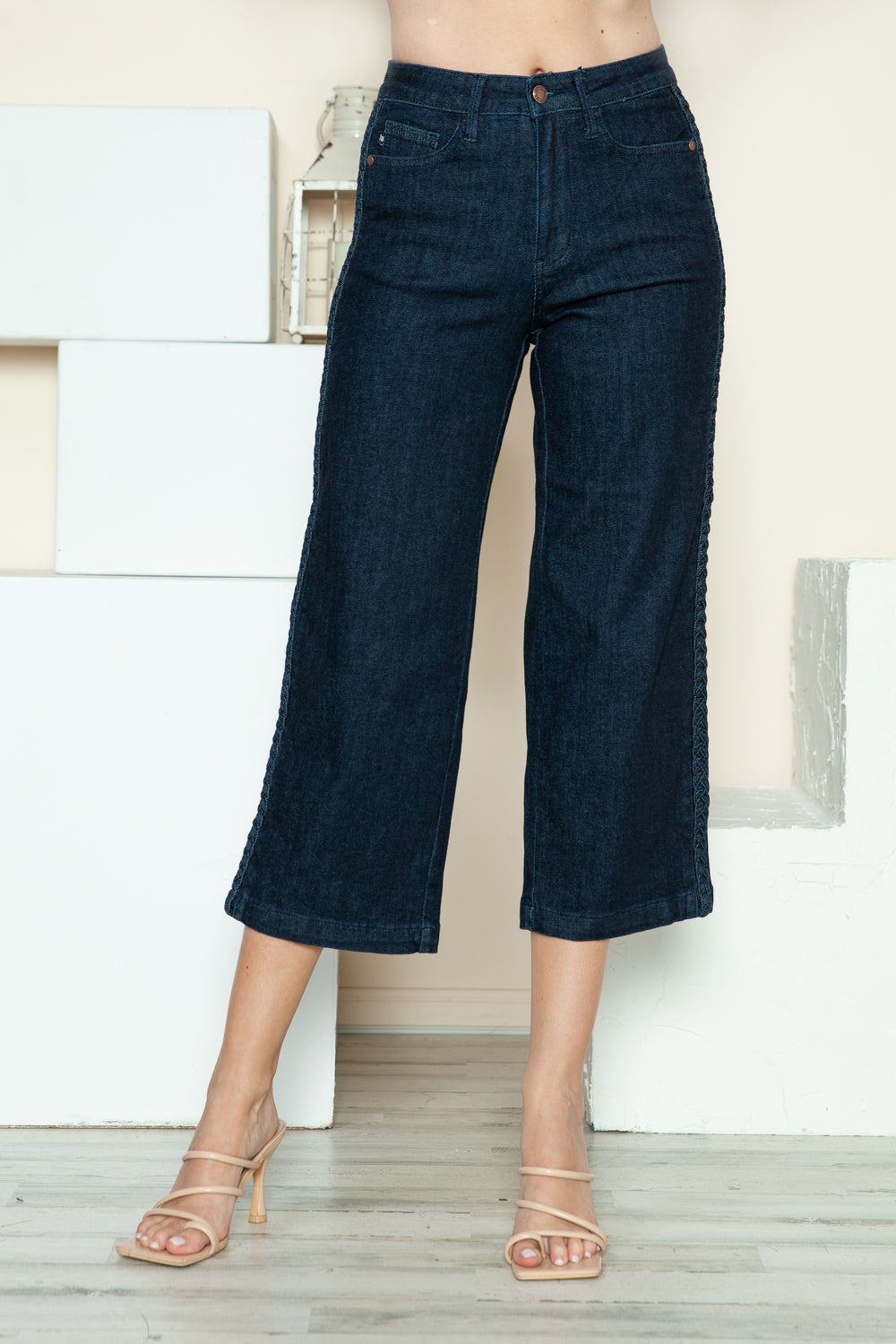 Person wearing Judy Blue Full Size Side Seam Braid Detail Crop Wide Leg Jeans and beige strappy heels, standing on a wooden floor with a white background.