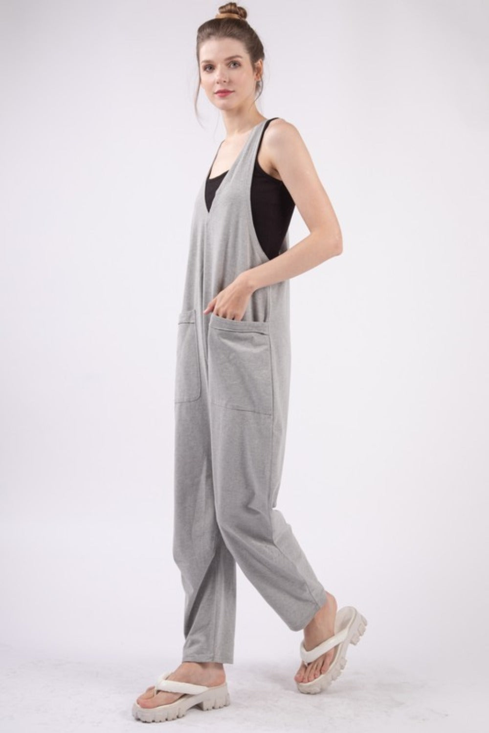 A person wearing the VERY J Plunge Sleeveless Jumpsuit with Pockets in gray and white sandals stands against a plain white background.