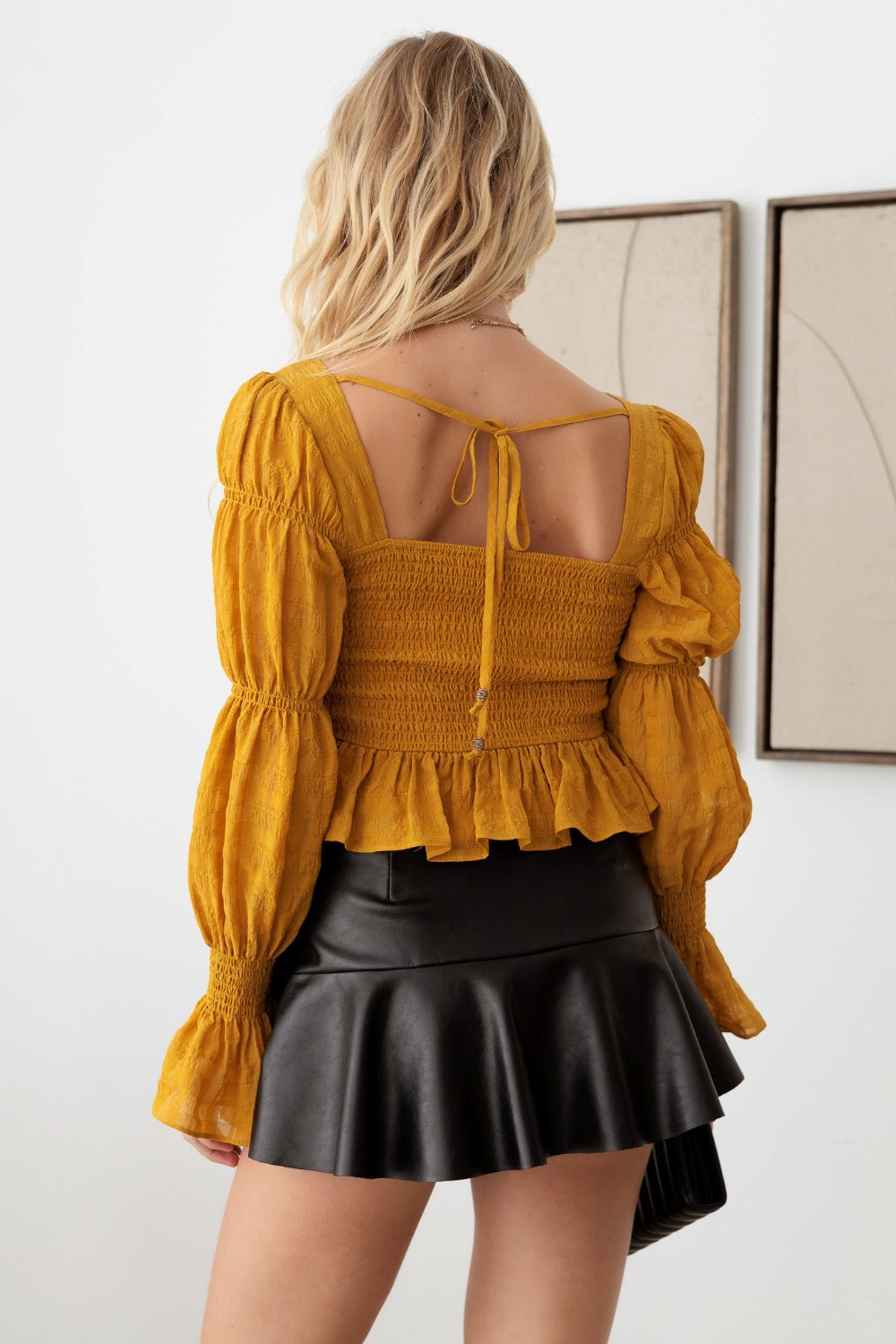 A woman with long blonde hair is wearing the Mustard Seed Boho Shirred Smocked Peplum Long Sleeve Top, a black skirt, and holding a black purse. She is standing in front of a white wall adorned with framed art.