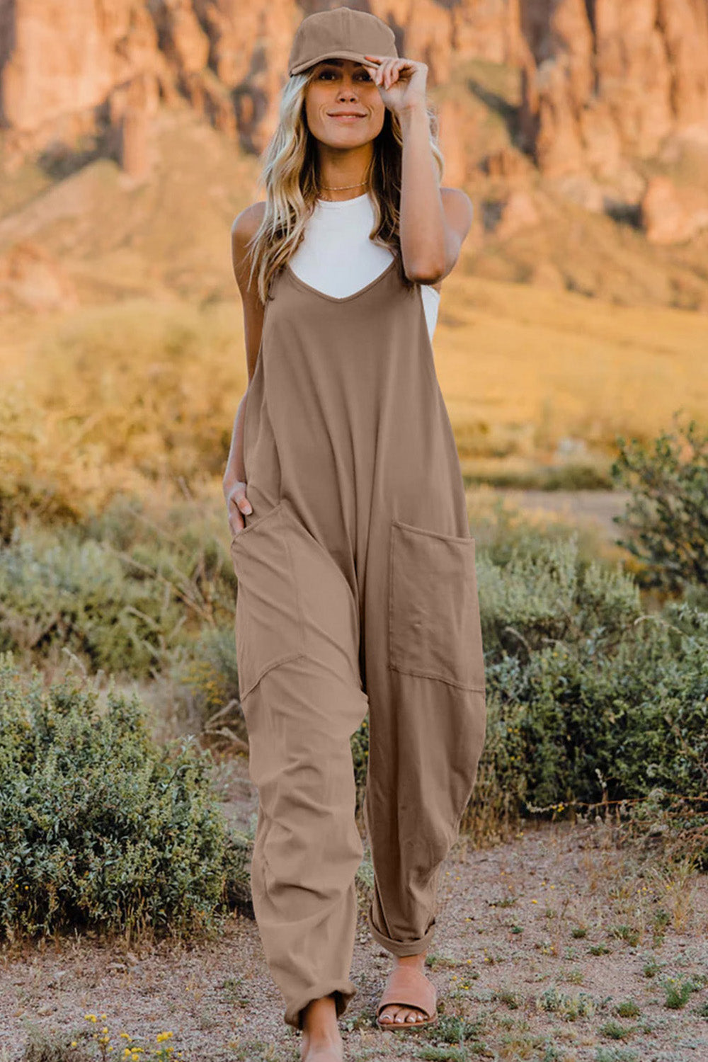 Person wearing a Double Take Full Size V-Neck Sleeveless Jumpsuit with Pockets and sandals, walking outdoors on a dirt trail with bushes in the background.