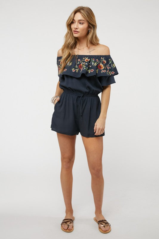 Wearing the stylish Floral Embroidered Off Shoulder Romper, a woman poses gracefully, showcasing its ruffle detail and making it an ideal addition to any summer wardrobe.