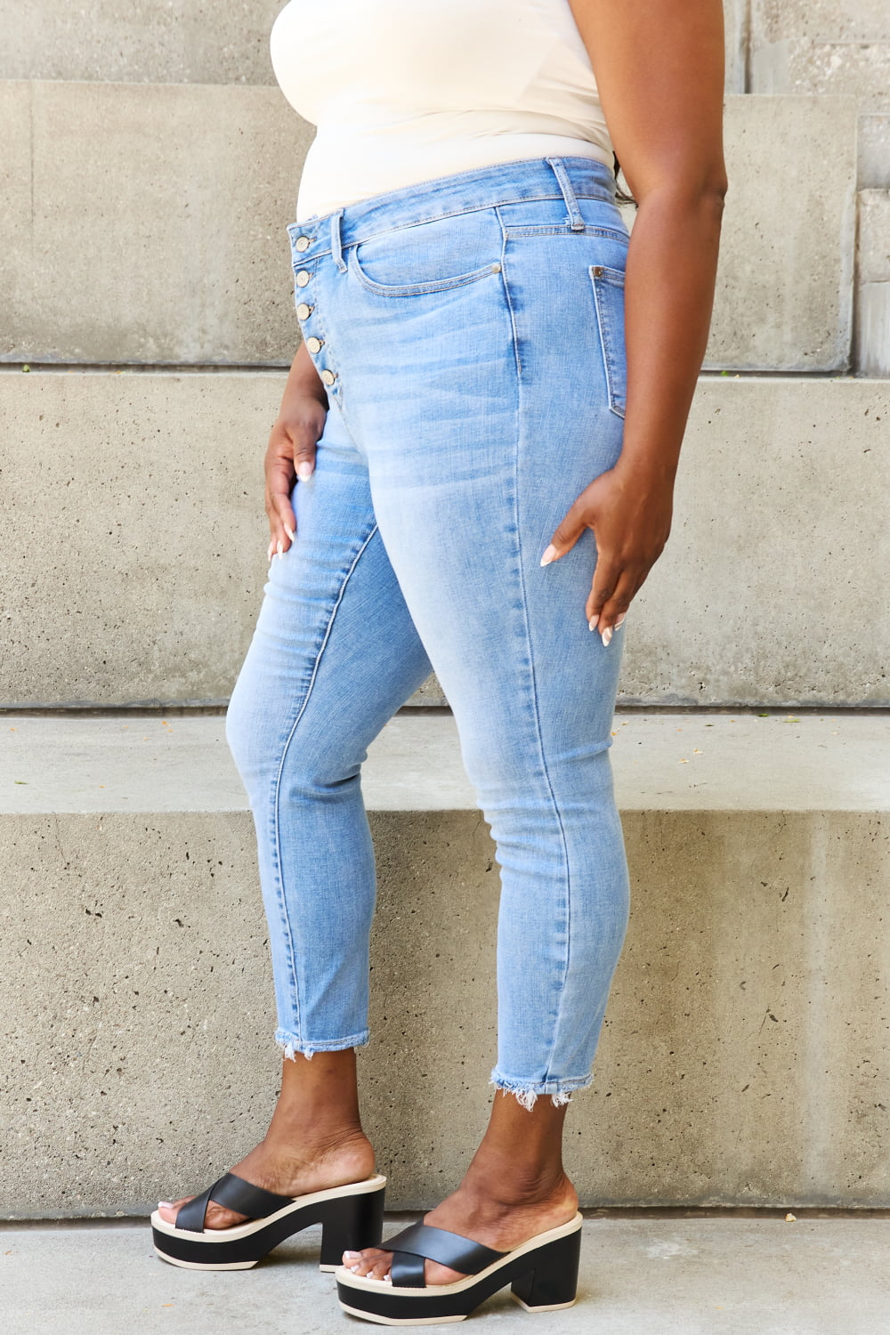 A person in Judy Blue Full Size Button Fly Raw Hem Jeans stands on concrete steps, showcasing high-waisted light blue denim with buttons and raw hems, blending style and comfort effortlessly.