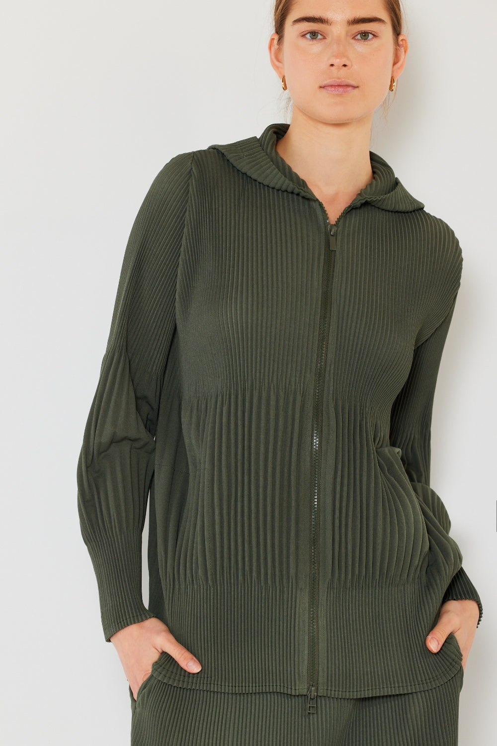 Individual wearing a dark green, long-sleeve pleated jacket with hands in pockets, highlighting its versatile unisex fit against a plain background.