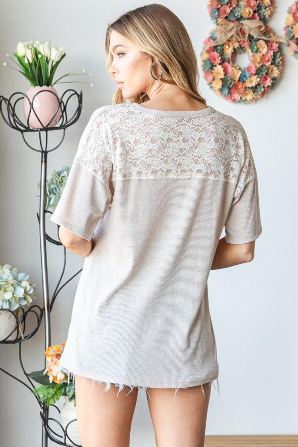 A woman wearing the Heimish Full Size Lace Front Pocket Drop Shoulder Top stands indoors near a decorative metal stand adorned with flowers.