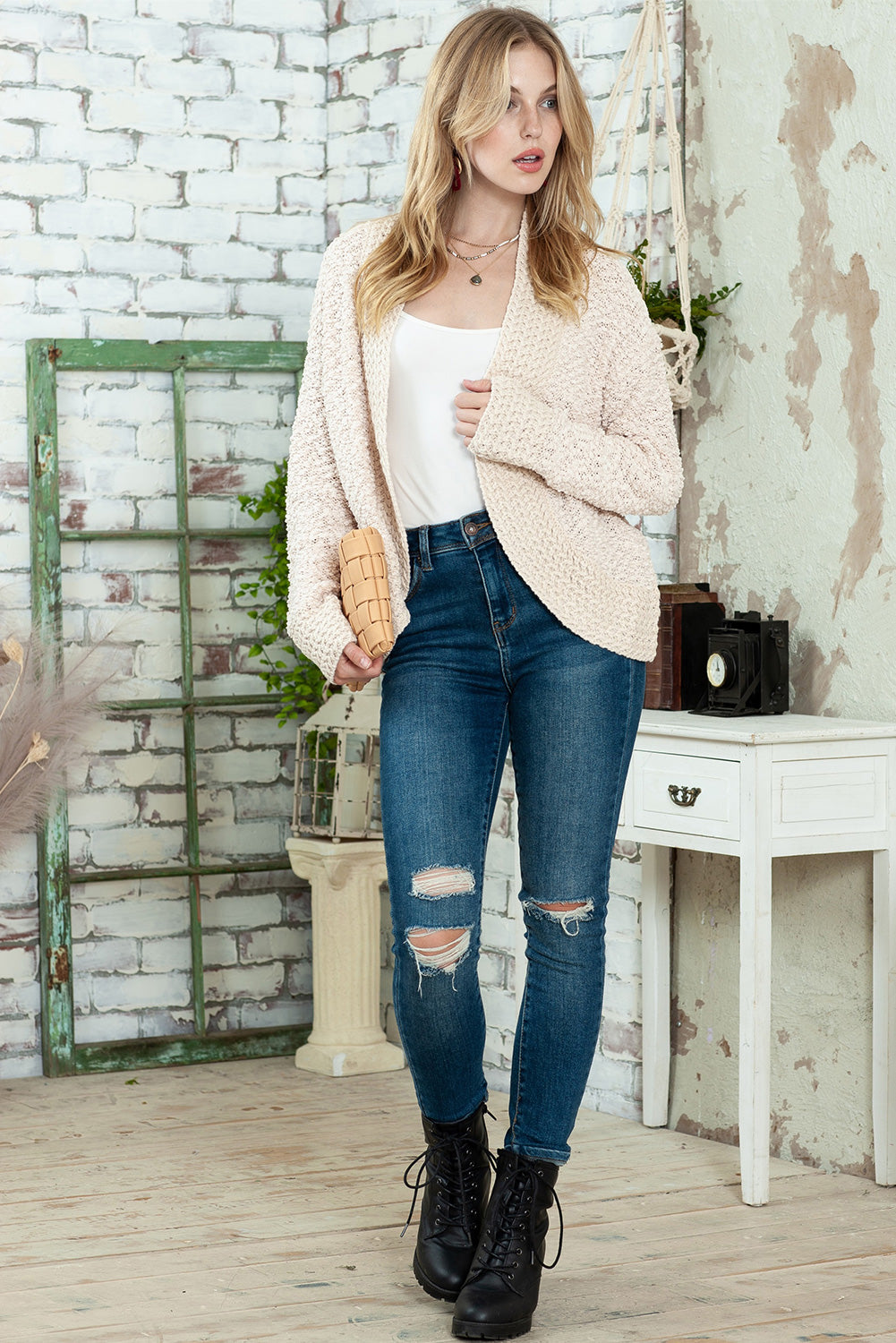 A woman in an Apricot Popcorn Knit Open Front Cardigan and jeans stands against a brick wall, looking to the side with a slight smile.