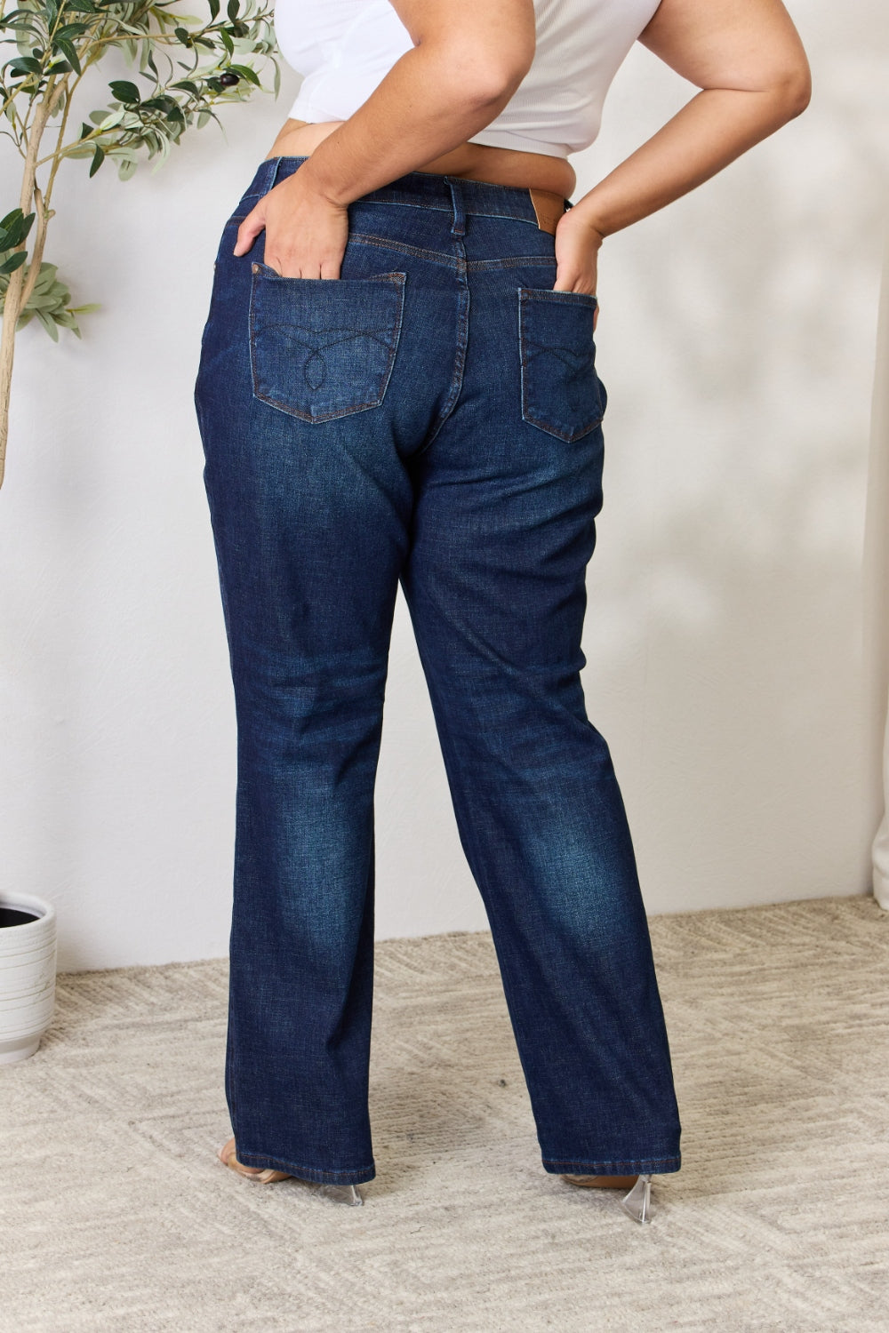 A person stands with hands in pockets, wearing the Judy Blue Full Size Button-Fly Straight Jeans, known for their high-waisted dark blue design crafted from premium lycra fabric. The jeans showcase button details and a straight leg cut, complemented by a white top and open-toed heels.