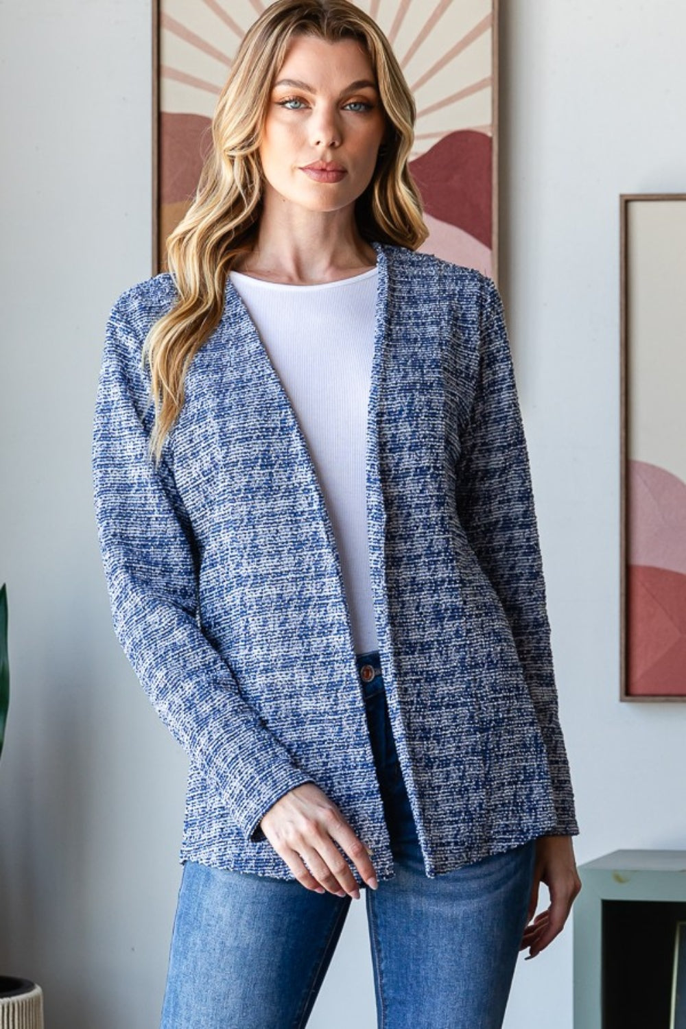 A woman with wavy blonde hair wears the Heimish Full Size Houndstooth Tweed Open Front Blazer in blue, paired with a white top and blue jeans. She stands in front of a wall adorned with framed art.