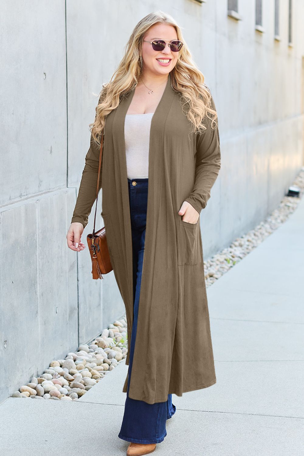 A woman stands against a concrete wall, wearing the Basic Bae Full Size Open Front Long Sleeve Cover Up made from rayon spandex, a white top, blue jeans in her perfect size, and brown shoes with her hands in her pockets. Machine wash cold for best results.