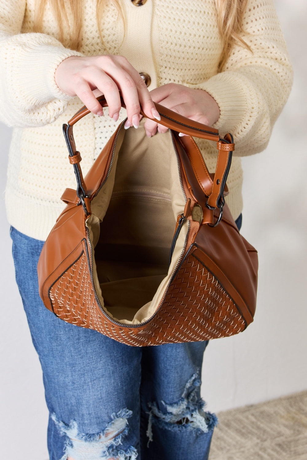 A person carries the SHOMICO Weaved Vegan Leather Handbag in a stylish brown hue, featuring a sleek crossbody strap, expertly paired with a cream sweater and jeans.