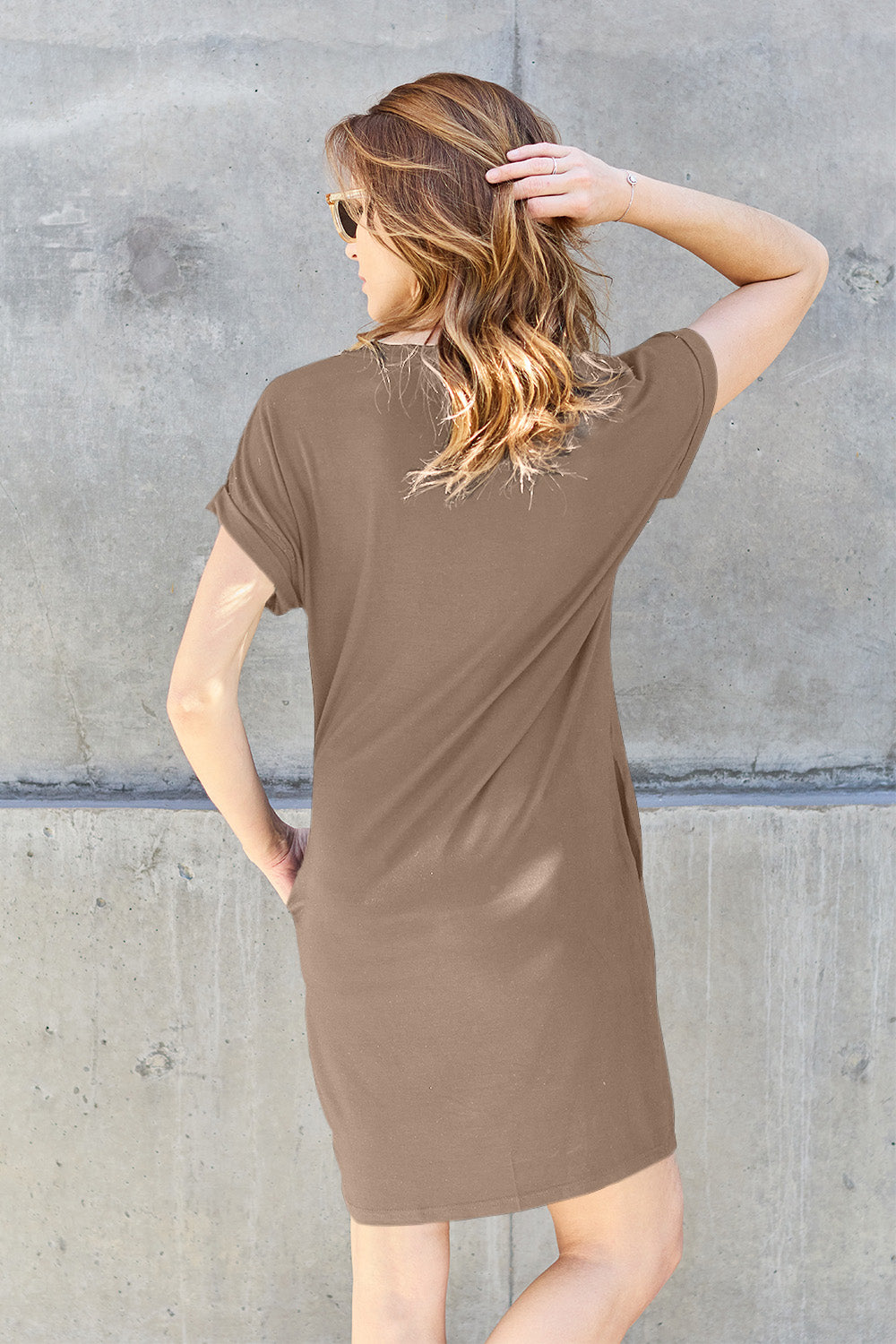 A woman wearing a dark olive-green Basic Bae Full Size Round Neck Short Sleeve Dress with Pockets and sunglasses stands against a concrete wall, holding a light-colored backpack.