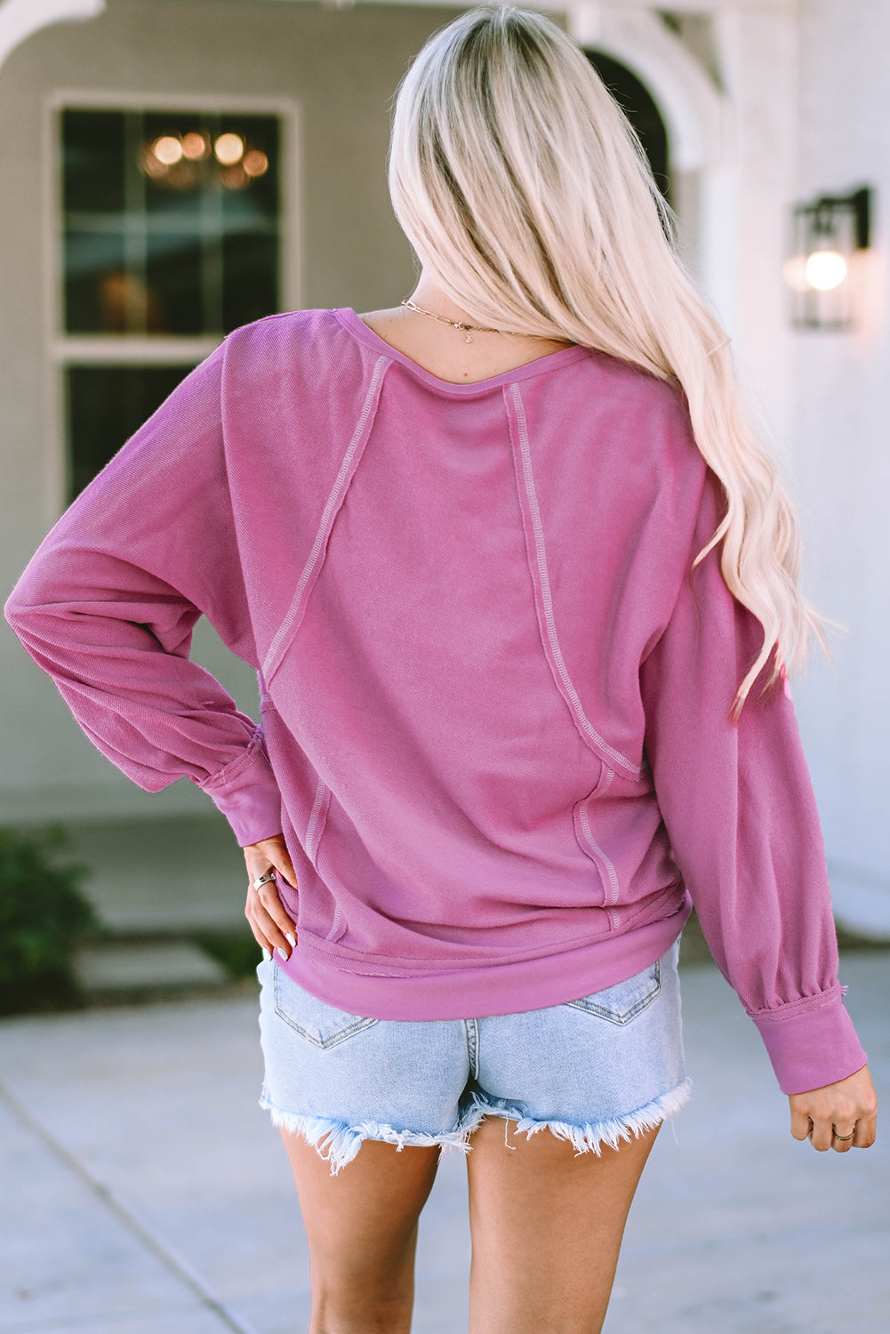 A person with long blonde hair is seen from the back wearing a Pink Exposed Seam Round Neck Terry Pullover and distressed denim shorts. The trendy look complements the casual outfit as they stand outdoors in front of a building with a window and a lantern.