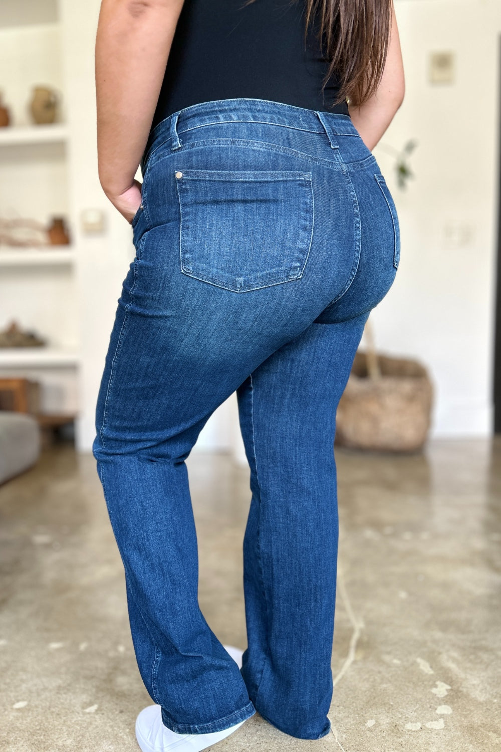 A person wearing a black top and Judy Blue Full Size Tummy Control Straight Jeans stands indoors with one hand on their hip. Their shoes are black with white soles. A plant and shelves are in the background.