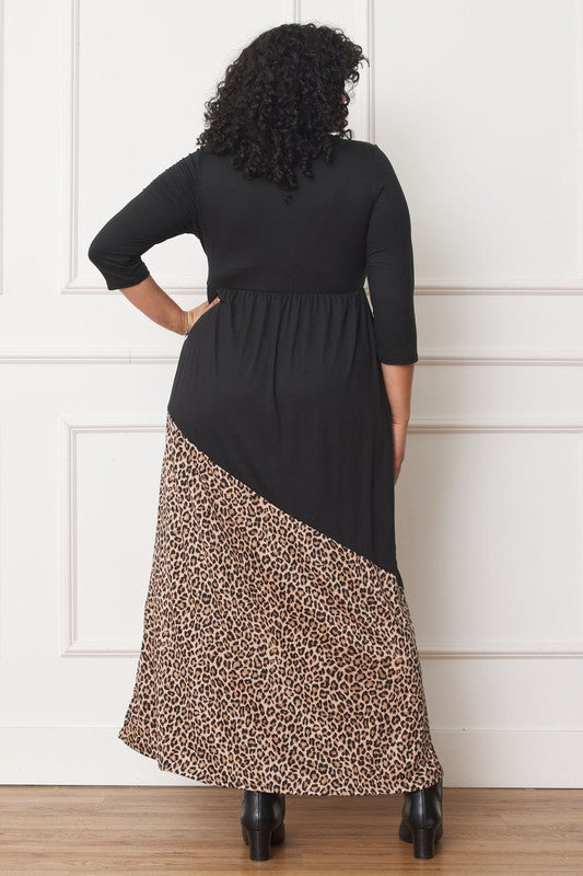 A woman with curly hair is wearing the Asymmetrical Leopard Accent Maxi Dress, featuring a long black design with a floral patterned section at the bottom. She stands against a white paneled wall, her hands tucked in her dress pockets, showcasing its elegant design.