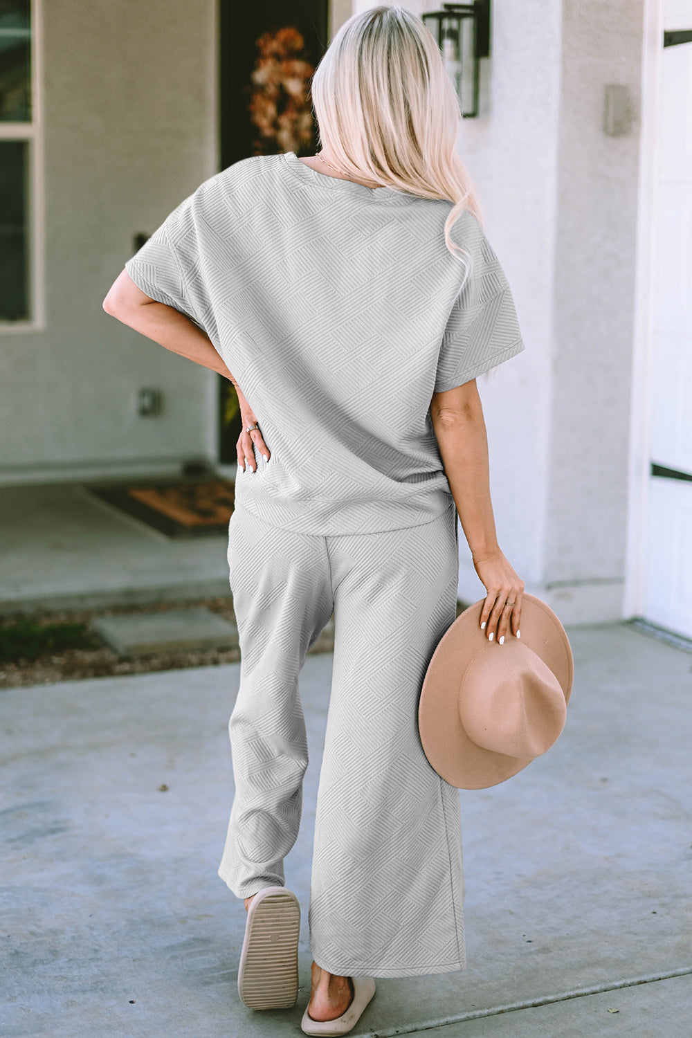 A woman with long hair smiles while standing outside a building, dressed in the Double Take Full Size Texture Short Sleeve Top and Pants Set, featuring a matching beige two-piece ensemble. She wears slides and holds a hat in her right hand. Her outfit, slightly stretchy for added comfort, is perfect for machine wash cold.