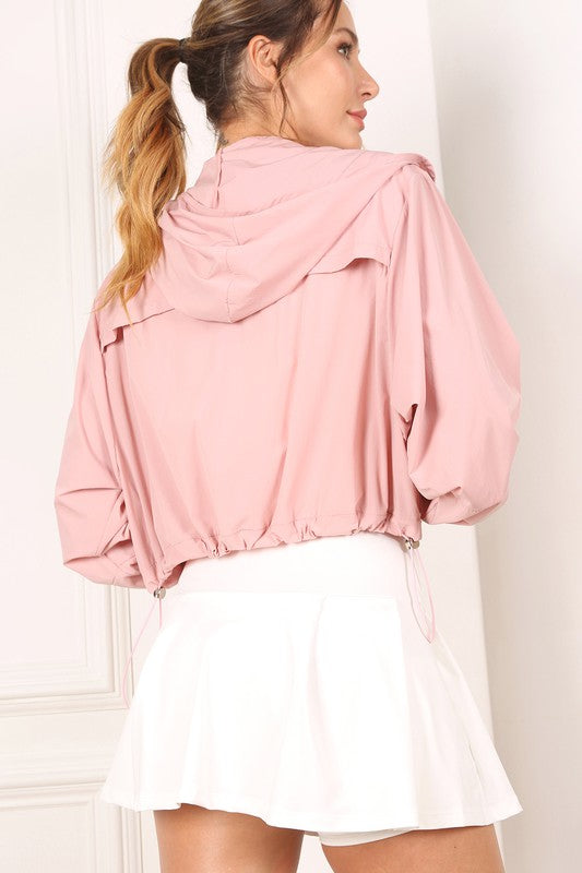 A woman wearing a pink wind breaker over a white top stands against a white wall.