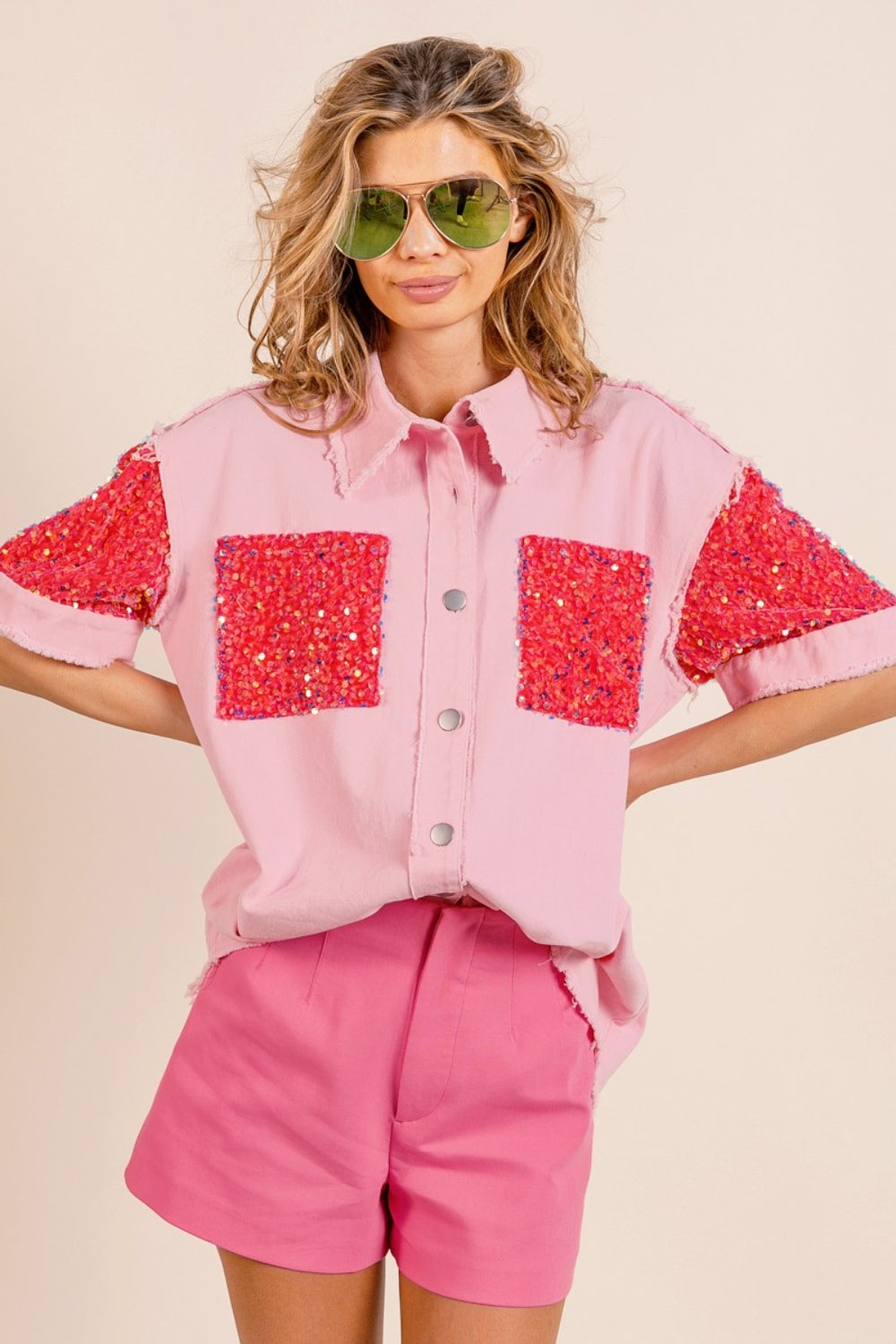 A woman poses in the BiBi Sequin Detail Raw Hem Short Sleeve Shirt, a pink button-up with red sequin detail pockets, paired with matching pink shorts and reflective aviator sunglasses.