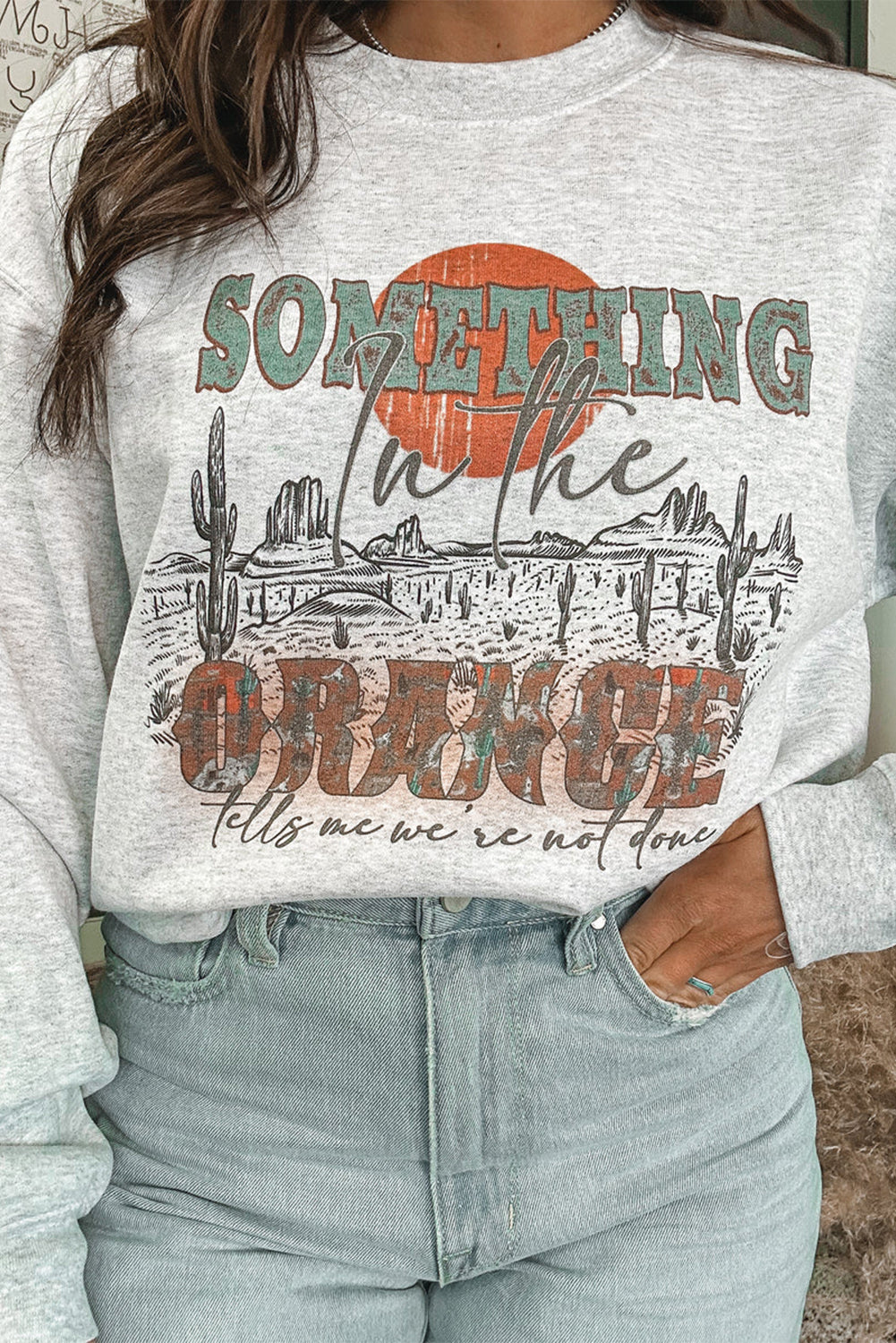 A person with long blonde hair is seen from behind, wearing denim shorts and a Gray SOMETHING ORANGE Graphic Relaxed Sweatshirt featuring a desert sunrise print.