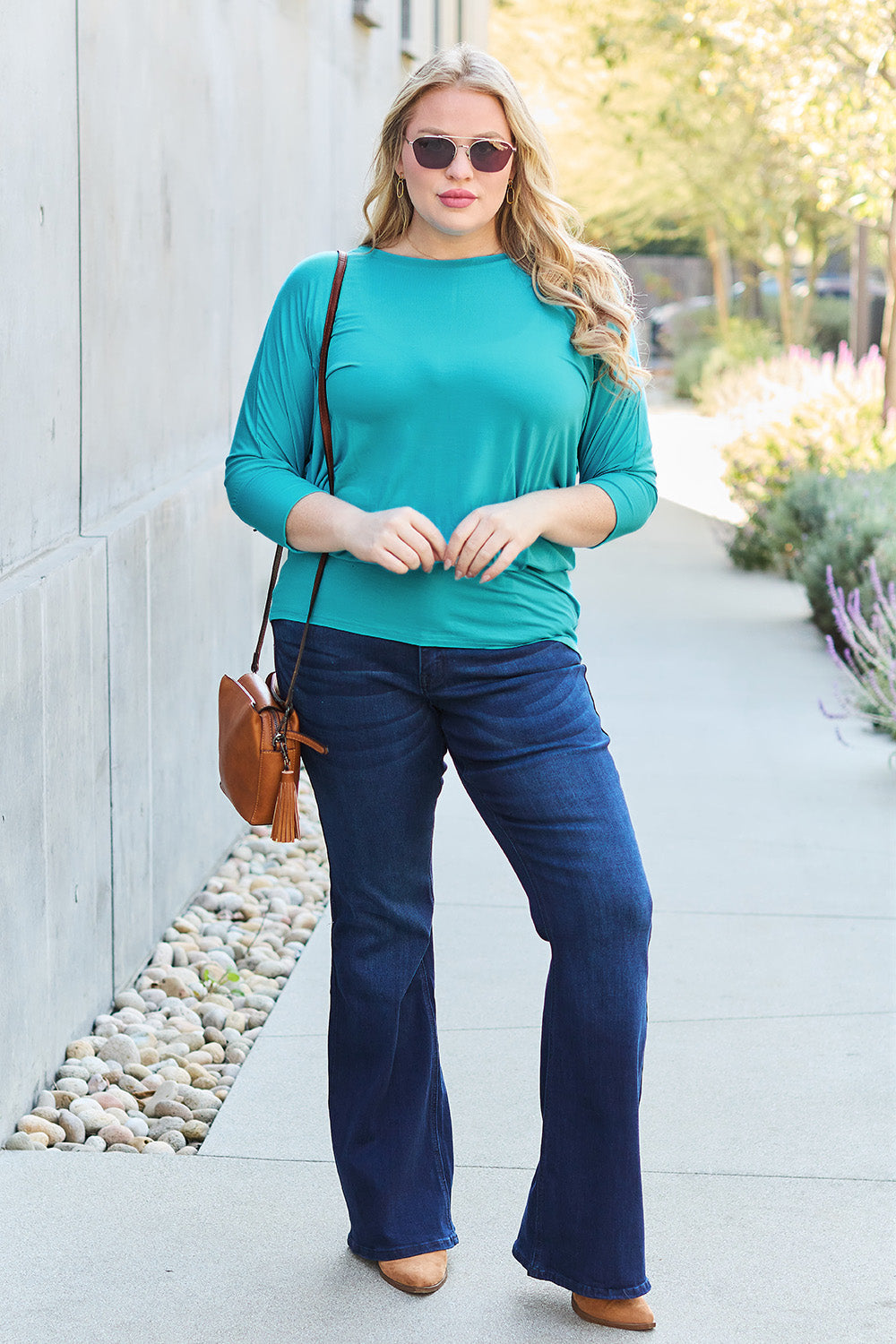 A woman in a slightly stretchy, brown Basic Bae Full Size Round Neck Batwing Sleeve top and blue jeans stands against a concrete wall, holding a brown handbag.