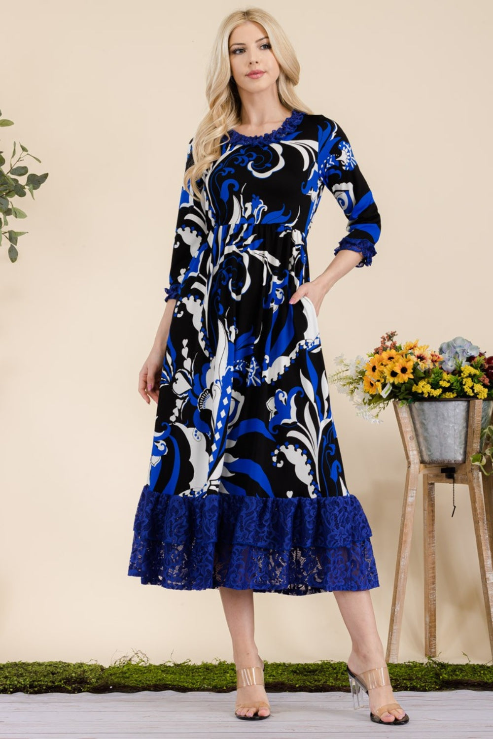 A woman wearing the Celeste Full Size Paisley Print Lace Ruffled Midi Dress stands near a wooden table adorned with a bucket of flowers.