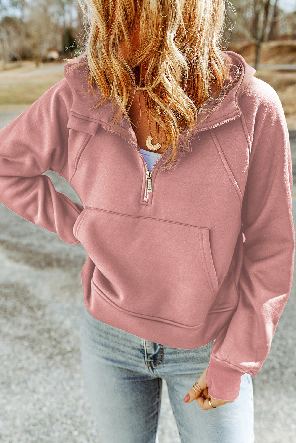 A person with wavy blonde hair is walking outside, sporting a Pink Quarter Zip Kangaroo Pocket Hoodie and light blue distressed jeans. The background includes a road and trees.