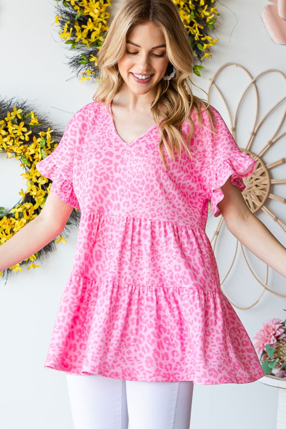 A person wearing a Heimish Full Size Leopard Ruffle Trim Short Sleeve Top stands in front of a floral wreath display.