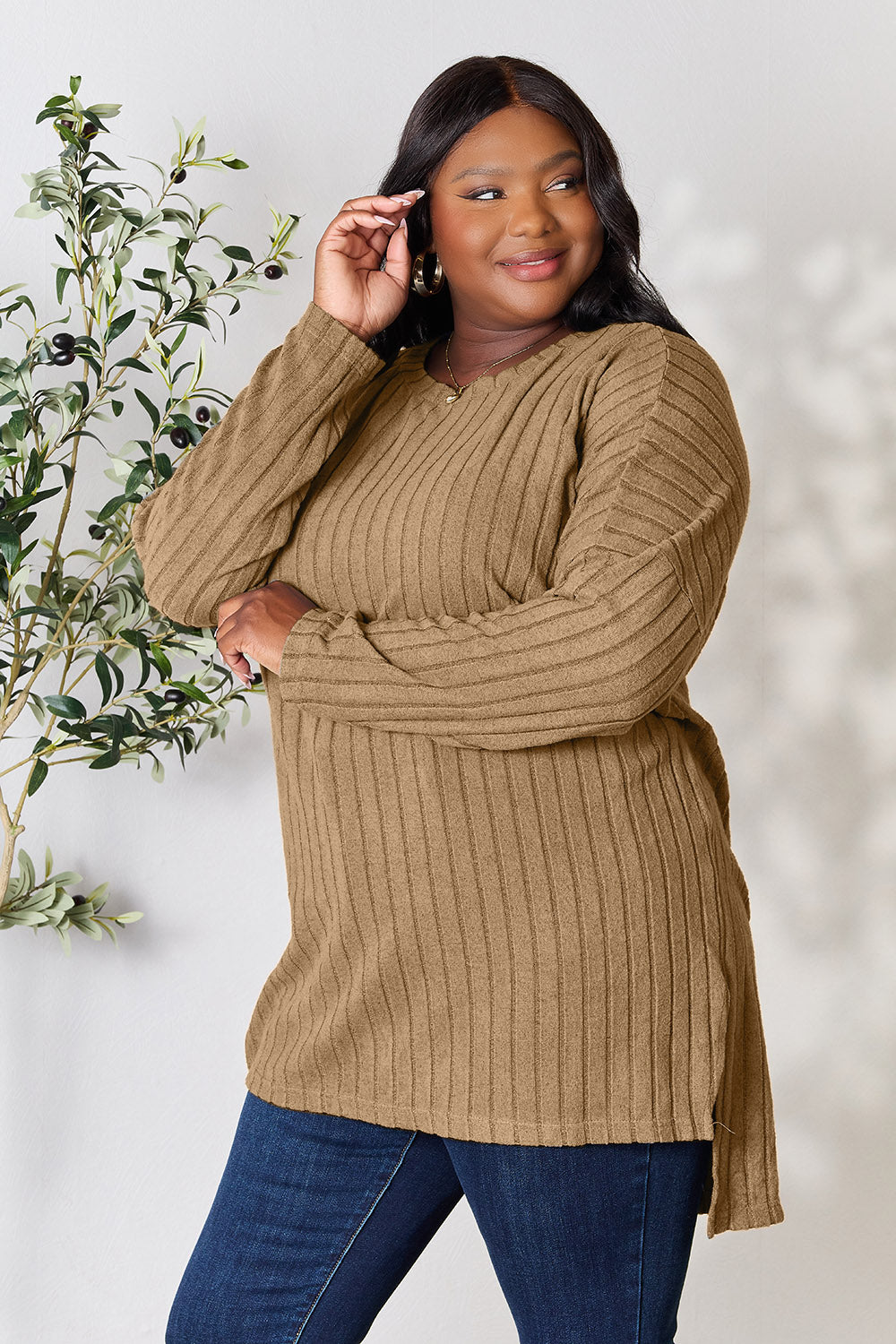 A person with shoulder-length hair smiles while wearing a beige ribbed sweater called the Basic Bae Full Size Ribbed Round Neck Long Sleeve Slit Top, along with matching beige pants, showcasing a basic style. They stand next to a green plant.