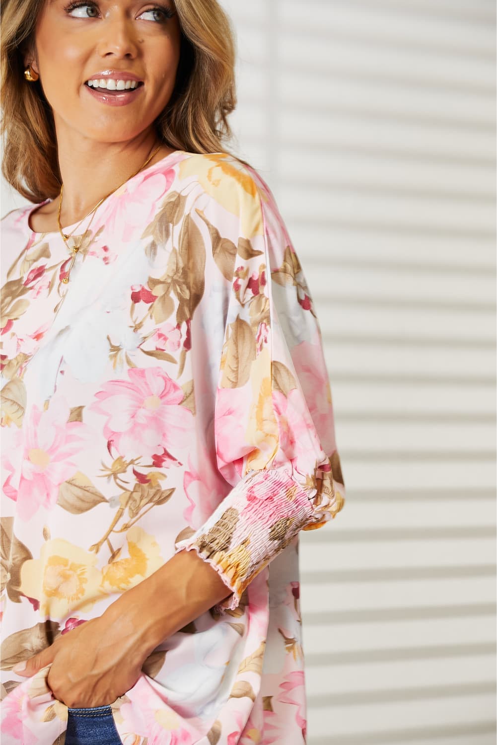 A woman in a Double Take Floral Round Neck Three-Quarter Sleeve Top and jeans stands smiling in a brightly lit room with a white background and drawers on the side.