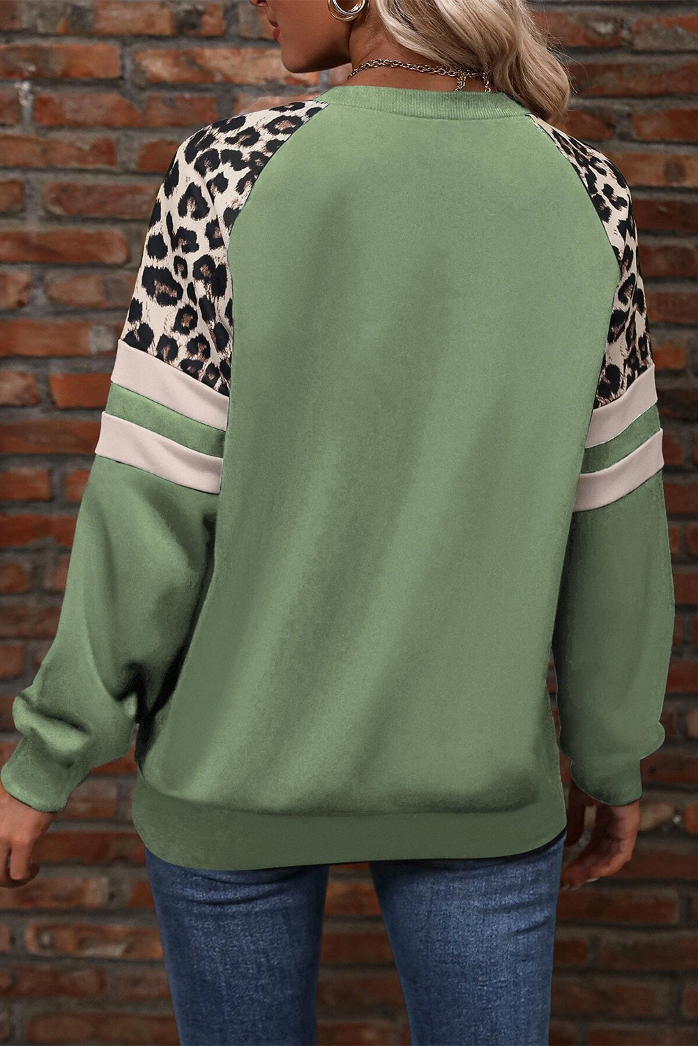 A person wearing a Grass Green Leopard Print Colorblock Raglan Sleeve Sweatshirt stands in front of a brick wall.