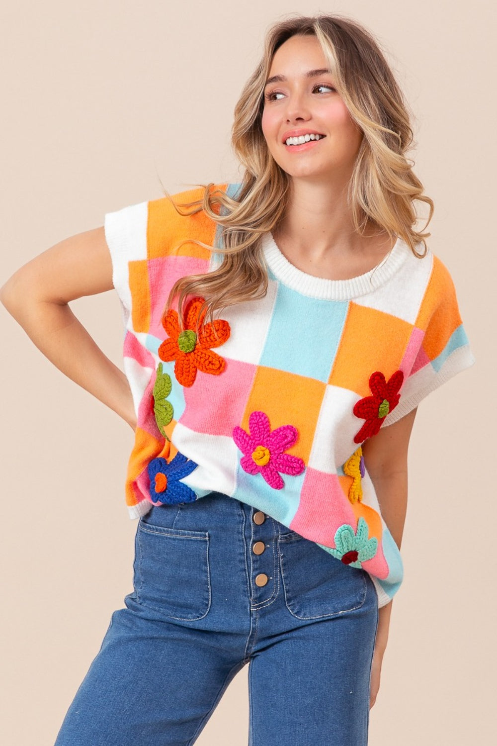 A woman is wearing the stylish BiBi Flower Patch Checkered Sweater Vest paired with high-waisted jeans. She has wavy blonde hair and a radiant smile while posing against a plain beige background. This trendy fall wardrobe piece adds a touch of warmth and whimsy to any outfit.