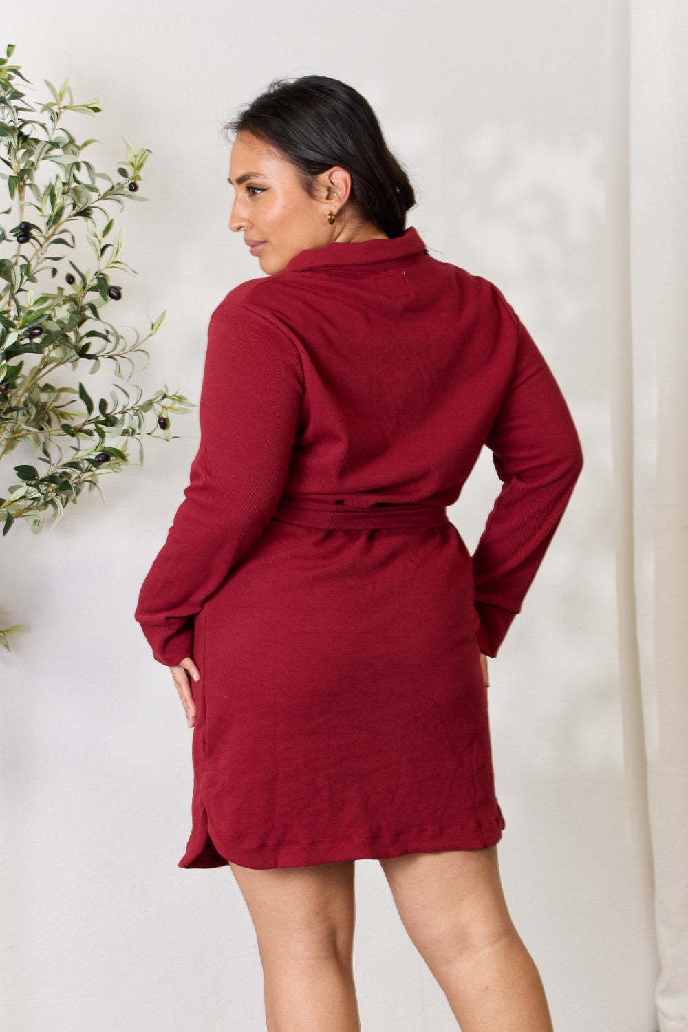 A woman wearing the Culture Code Full Size Tie Front Half Zip Long Sleeve Shirt Dress stands in front of a plain background next to a potted plant.