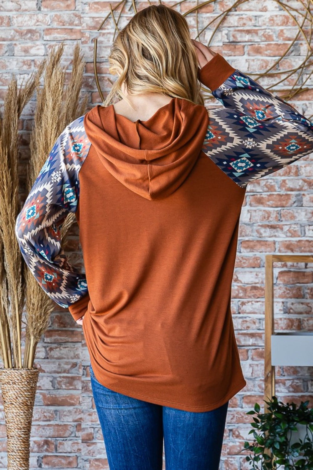 A woman in a Heimish Full Size Half Button Printed Long Sleeve Hooded Top poses casually in front of a brick wall, holding the hood with one hand. She is wearing blue jeans and standing near dried plants, showcasing the unique print design that adds an on-trend touch to her outfit.