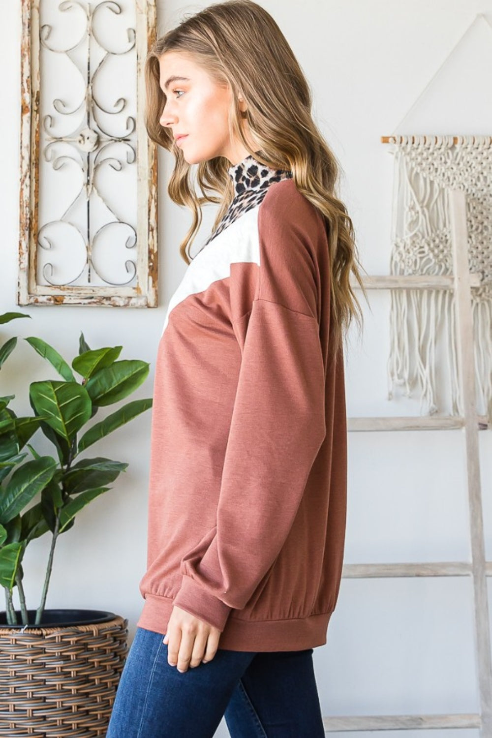 A woman wearing the stylish Heimish Full Size Leopard Quarter Zip Mock Neck Sweatshirt stands indoors near a lush plant and an intricate macrame wall hanging.