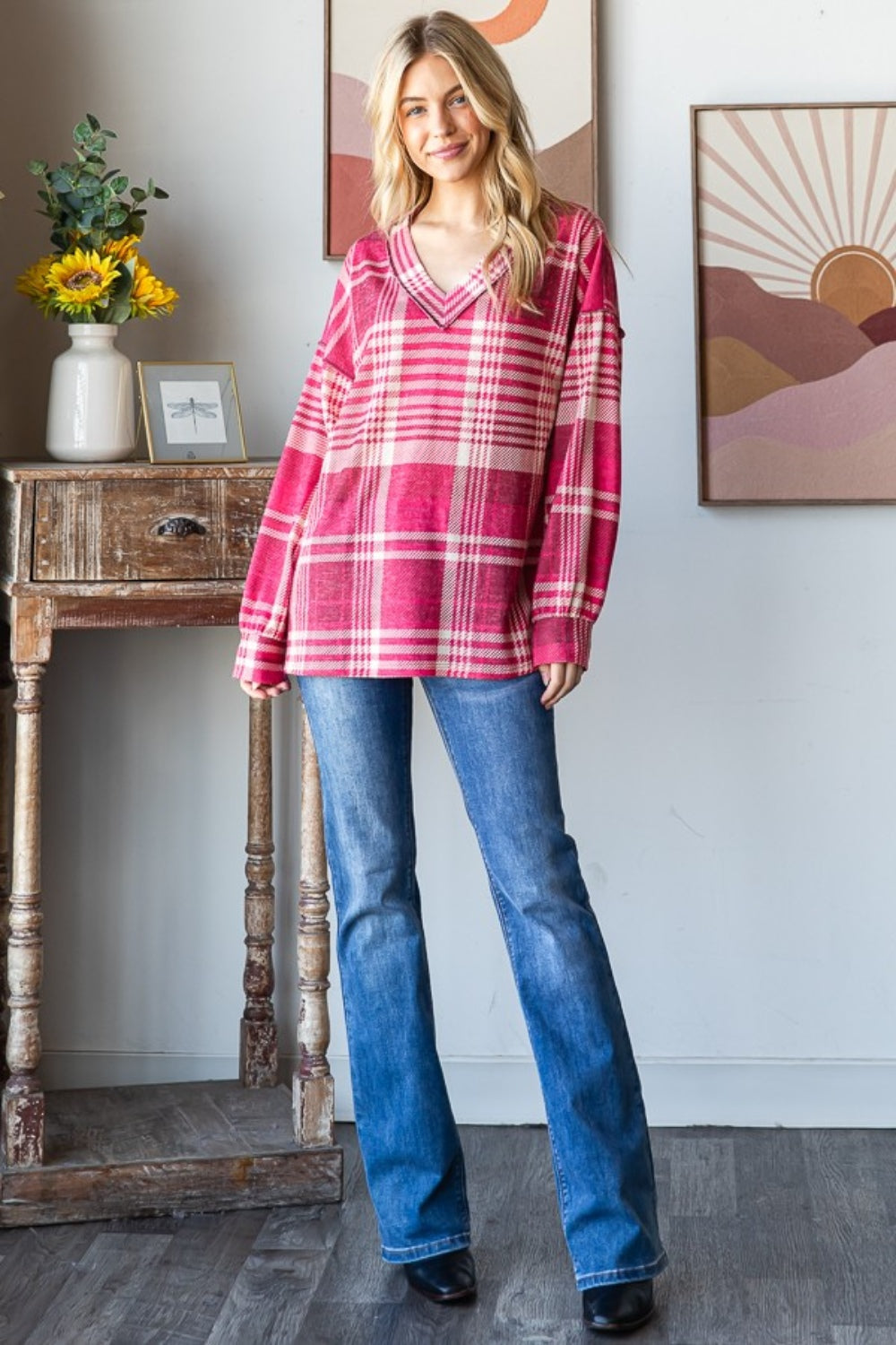 A person with long blond hair smiles while wearing the Heimish Plaid Drop Shoulder V-Neck T-Shirt in red and white and a pair of blue jeans, leaning against a wooden table with framed pictures in the background.