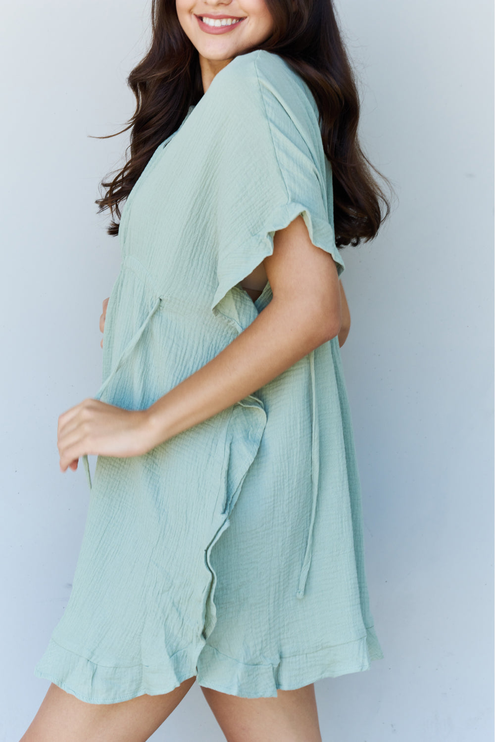 A woman is wearing the Ninexis Out Of Time Full Size Ruffle Hem Dress with Drawstring Waistband in Light Sage. Her long, dark hair falls gracefully as she smiles and looks to her left, standing against a plain background.