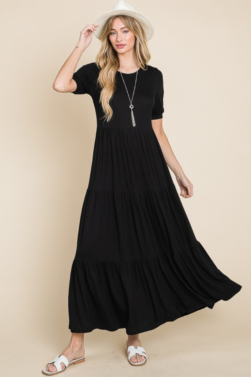 A person wearing the BOMBOM Short Sleeve Tiered Maxi Dress in sleek black stands against a plain beige background, exuding comfort with style. With one hand on their hip and donning white sandals, they effortlessly showcase this versatile piece.