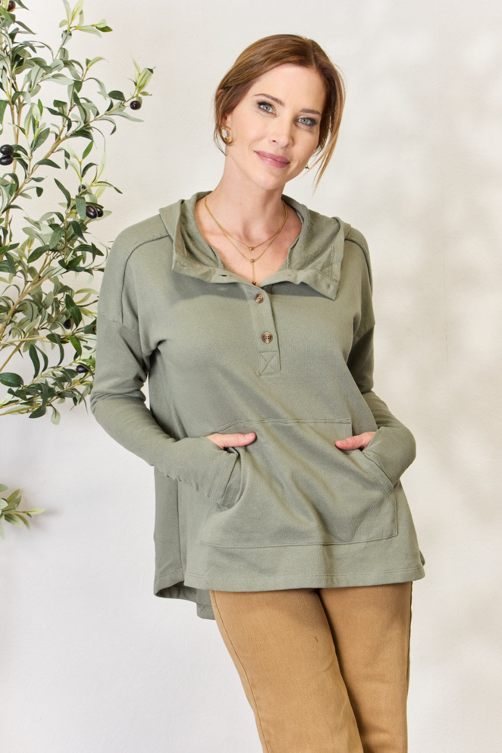 A person stands with hands in the pockets of their light green Culture Code Full Size Half Button Hoodie and brown pants. A plant is visible on the left side of the image.