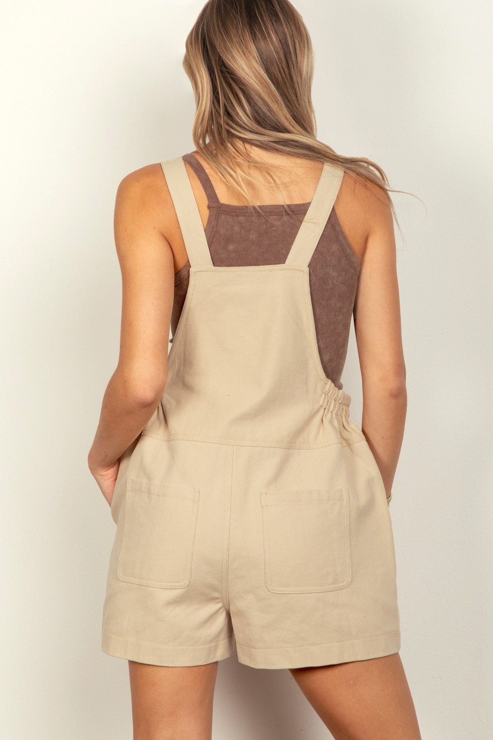 A woman wearing the VERY J Adjustable Suspender Overalls with Pockets in beige and a brown tank top smiles while touching her hair with one hand and has her other hand in a pocket.