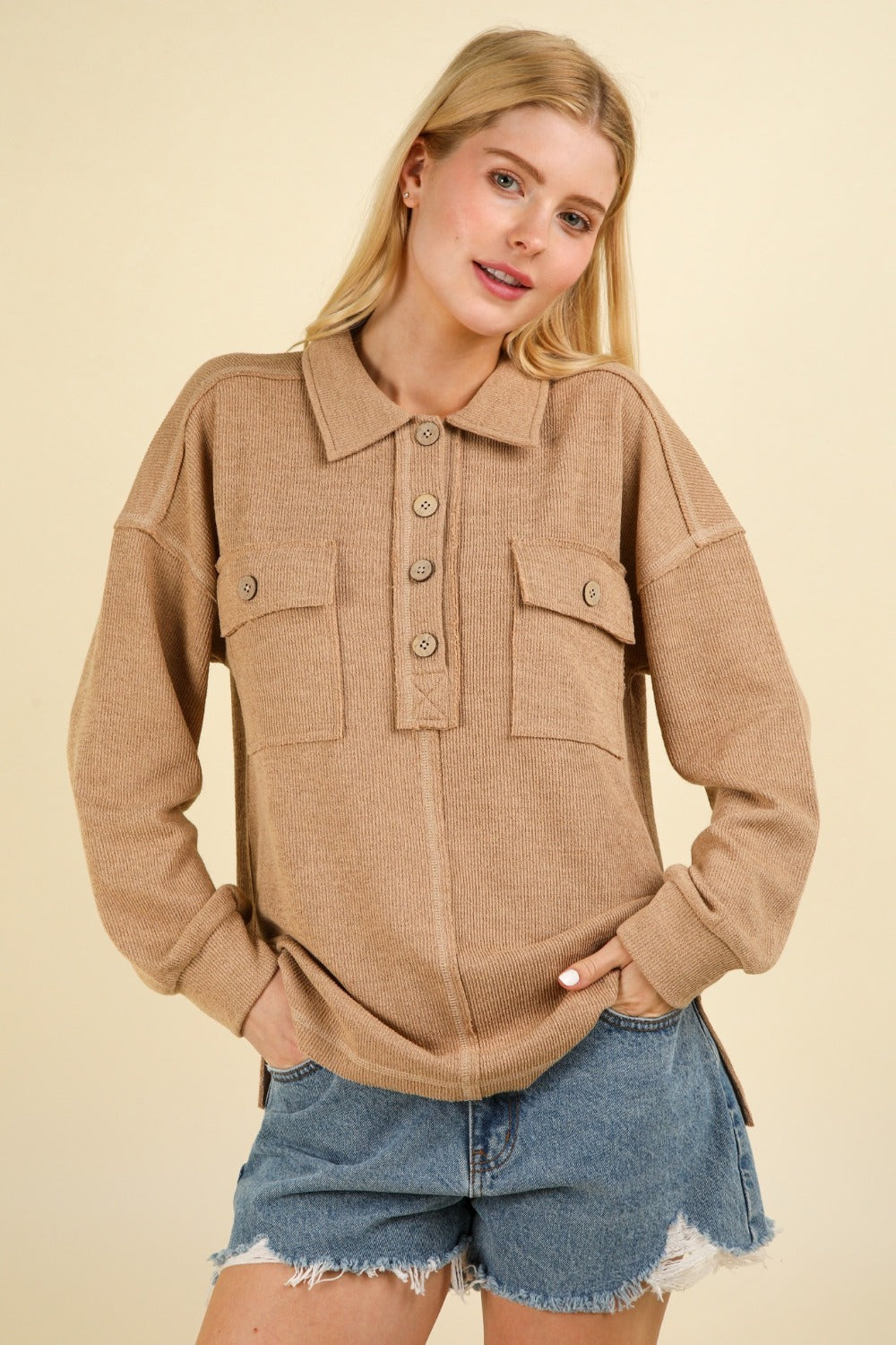 A woman with blonde hair wears the VERY J Collared Half Button Knit Top with Pockets in beige and denim shorts. She stands in front of a neutral background with her hands in her pockets.