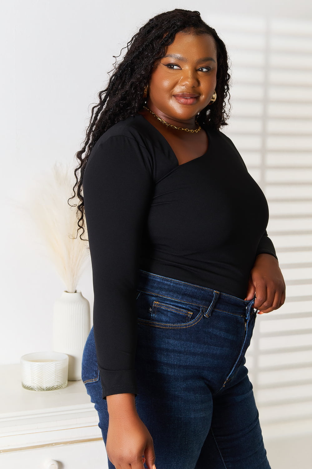 A woman with long hair is wearing a modern, stylish wardrobe featuring the Basic Bae Asymmetrical Neck Long Sleeve Top and jeans. She stands indoors in front of a white wall with shadow patterns.