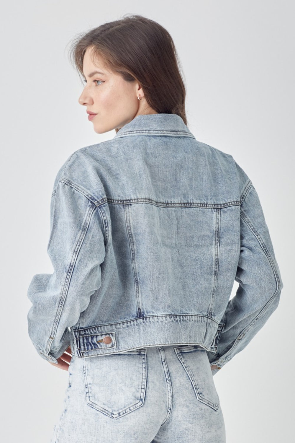 A woman wearing the RISEN Full Size Button Down Cropped Denim Jacket over a white top and distressed denim shorts stands against a plain background, showcasing versatile outerwear perfect for any casual occasion.