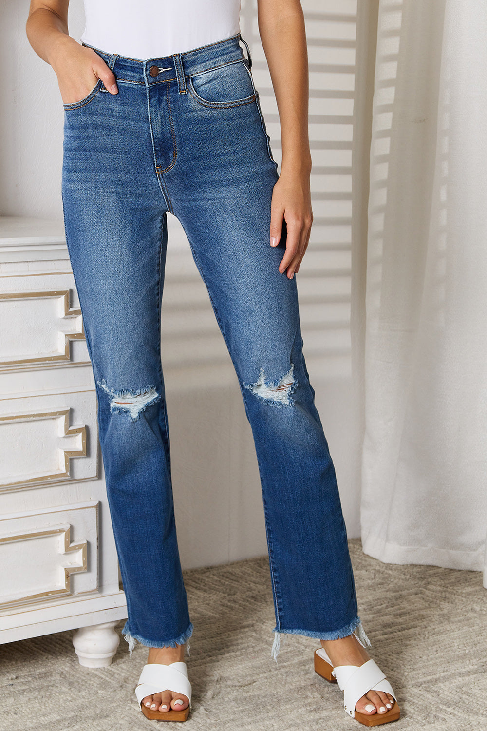 A person wearing Judy Blue Full Size Distressed Raw Hem Jeans, which are slightly stretchy and blue, along with a white top stands indoors near a white dresser.