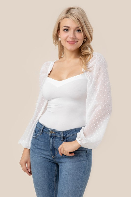 A person wearing the LS shirring sleeve bodysuit with Swiss dot mesh, featuring sheer, long puffed sleeves and a sweetheart neck, paired with light-colored jeans, stands against a plain background.