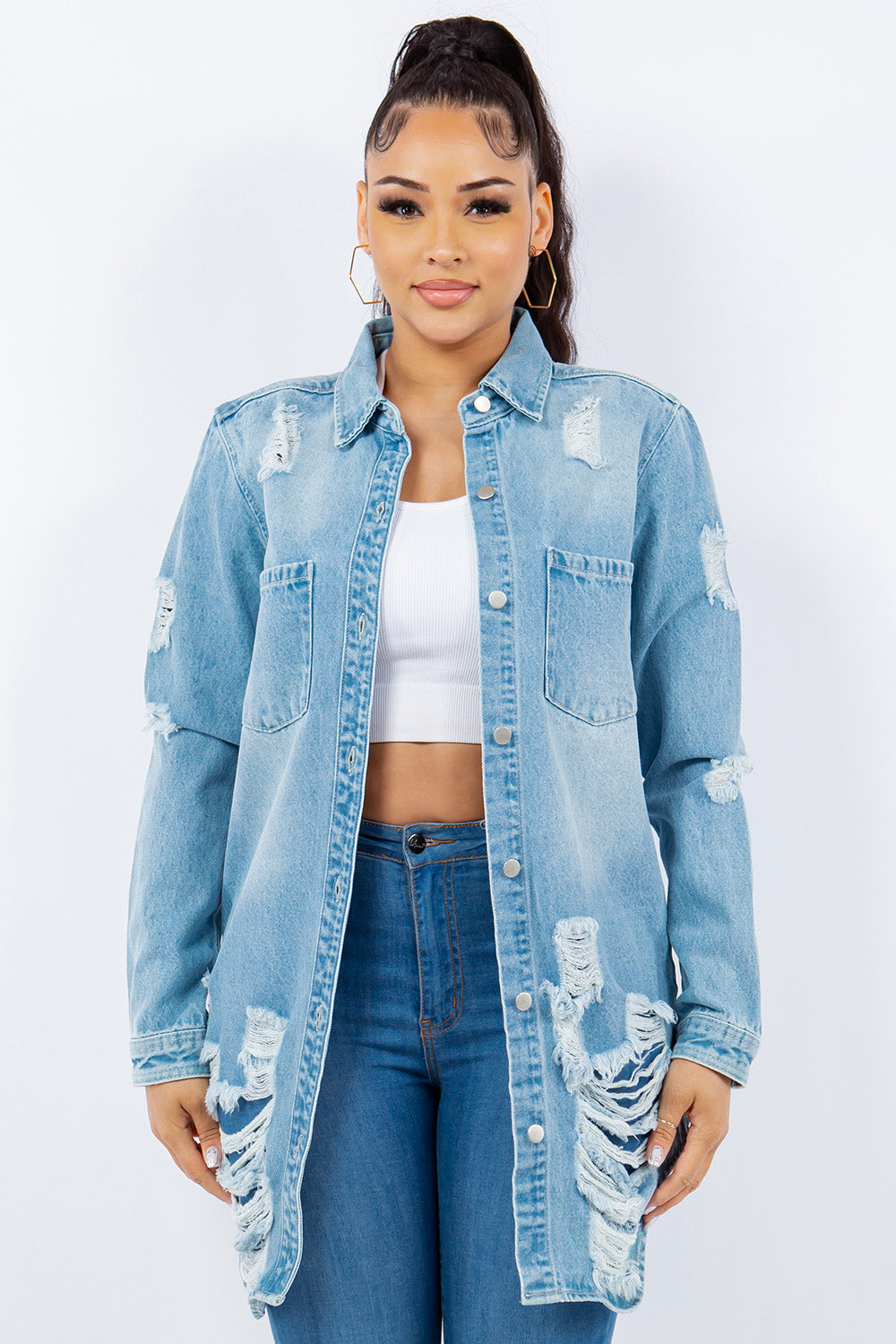 A woman with long hair is wearing an American Bazi Distressed Button Up Long Sleeve Denim Jacket over a white crop top and blue jeans. She is facing forward and standing against a plain white background.