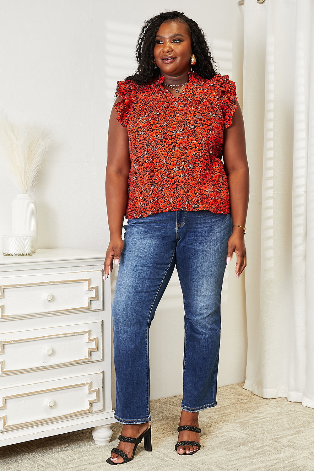 A woman wearing a chic Double Take Floral Flutter Sleeve Notched Neck Blouse and blue jeans stands smiling in a well-lit room with white walls and a curtain.