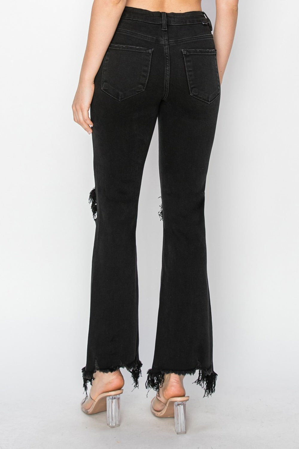 Rear view of a person wearing RISEN Full Size Distressed Raw Hem Jeans with Pockets in black, paired with clear-heeled sandals against a plain background.