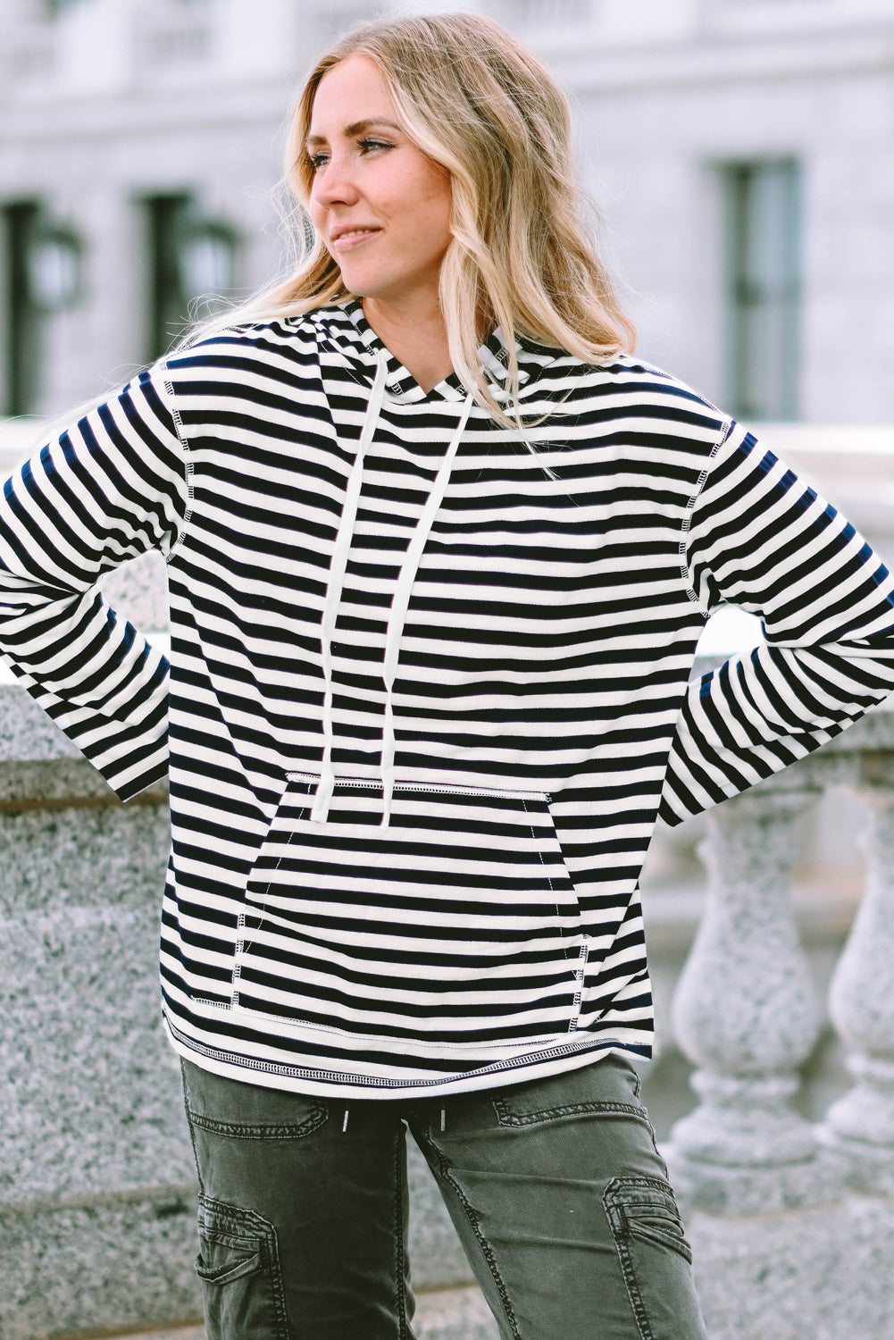 A person with long, blonde hair wearing a trendy Stripe Kangaroo Pocket Drawstring Hoodie with black and white stripes and dark green pants is standing outdoors, facing away from the camera.