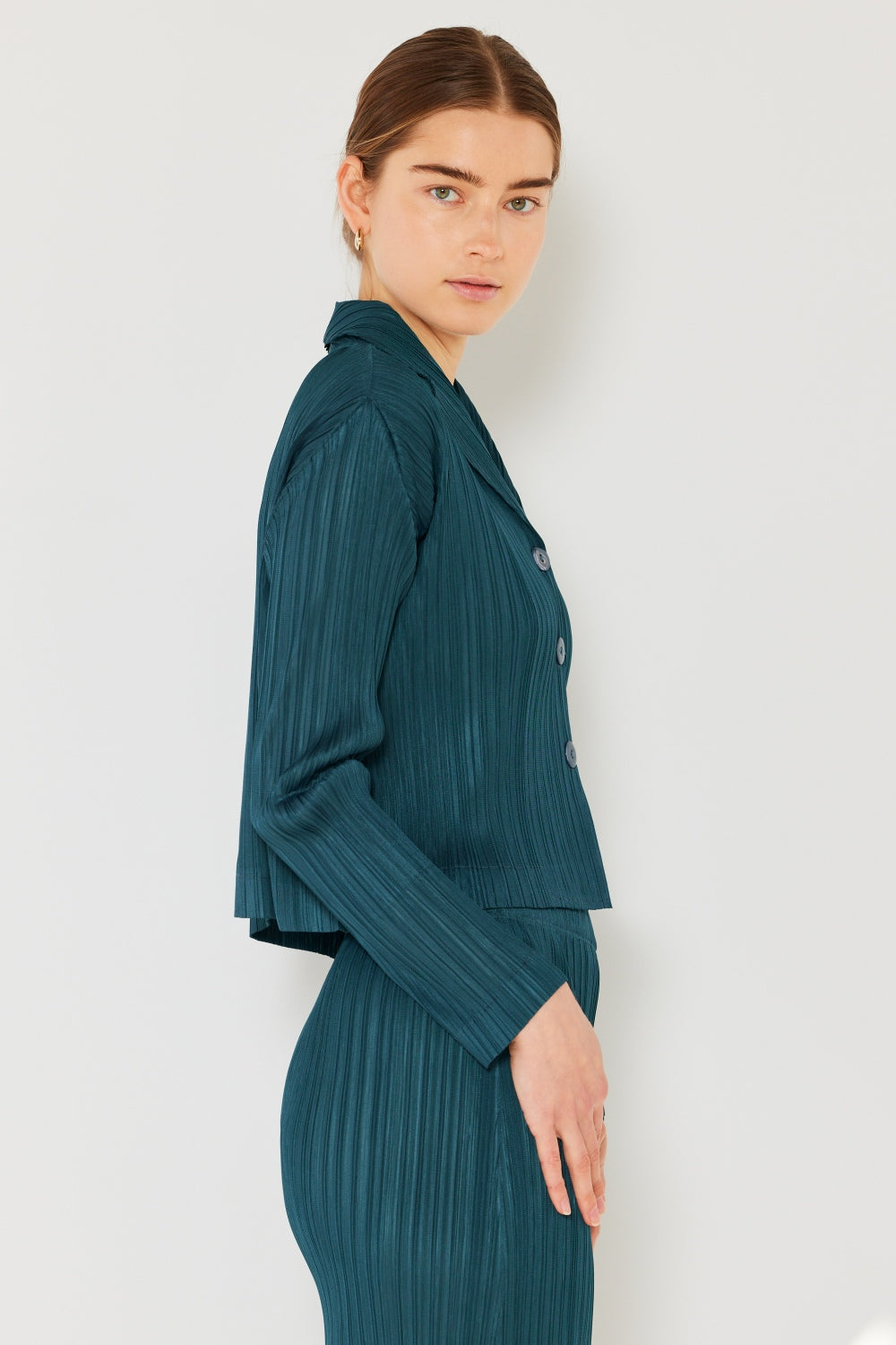 A person wearing the Marina West Swim Pleated Cropped Button Up Shirt in teal, complete with long sleeves and a matching skirt, stands against a light background, adding a modern touch to the classic pleats.